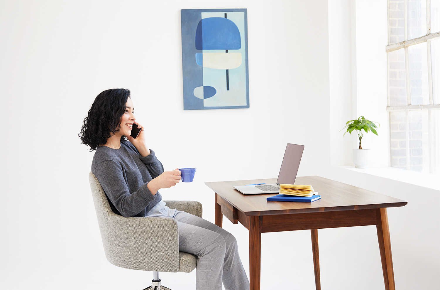 A woman speaking on her cell phone and holding a cup of coffee in front of a desk that has an open laptop.