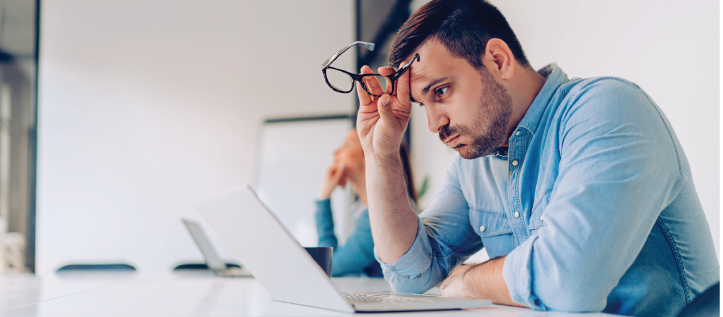 A stressed man holding his glasses while looking at his computer screen